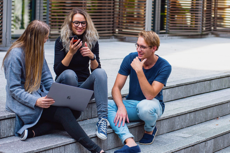 3 blide studenter som sitter med mobil og pc og snakker sammen utenfor skolen.